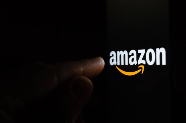 A hand is shown reaching towards a smartphone screen that displays the Amazon logo, which consists of the word "amazon" with an orange arrow forming a smile underneath the text, connecting the letters "a" and "z". The background is dark. - Cuponsy