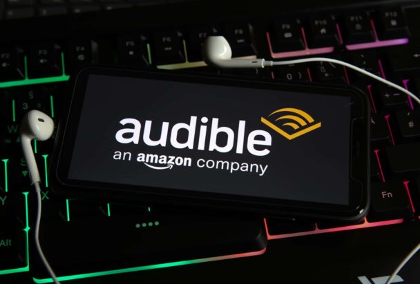 A smartphone displaying the Audible logo with the tagline "an Amazon company" rests on a colorful backlit keyboard. Earbuds are placed around the phone. - Cuponsy