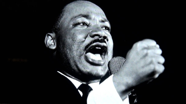 A black and white photo of a person wearing a suit and tie, speaking passionately into a microphone. The person's expressive face and raised hand convey a powerful and emotive speech. The background is dark, focusing attention on the speaker. - Cuponsy