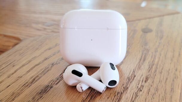 A pair of white wireless earbuds are placed on a wooden surface in front of their matching white charging case. The earbuds have black accents on the speaker and microphone areas. The background is a blurred indoor setting. - Cuponsy