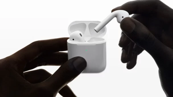 A person holds a wireless earbud above its open charging case with both hands. The case has a small green light indicating it's powered on. The background is white, making the earbud and charging case stand out prominently. - Cuponsy