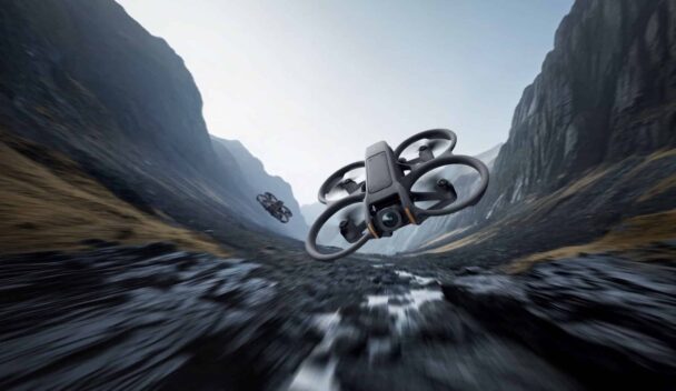 Two drones equipped with cameras fly through a rugged, narrow mountain valley with rocky terrain. The scene is set against a backdrop of steep cliff faces and a cloudy sky, creating a dynamic and adventurous atmosphere. - Cuponsy