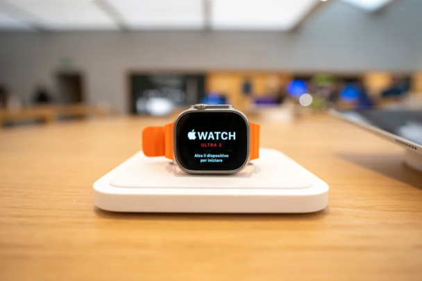 A close-up of an Apple Watch Ultra 2 on display in an Apple Store. The watch has an orange band and is placed on a white stand. The screen reads "Apple Watch Ultra 2" with instructions in Italian to lift the device to start. The background shows a blurred store interior. - Cuponsy