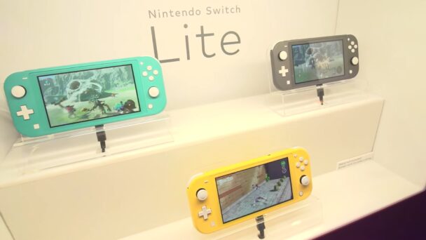 A display features three Nintendo Switch Lite consoles in turquoise, gray, and yellow colors mounted on stands. Each console shows different game scenes on their screens. The text "Nintendo Switch Lite" is visible above the consoles. - Cuponsy