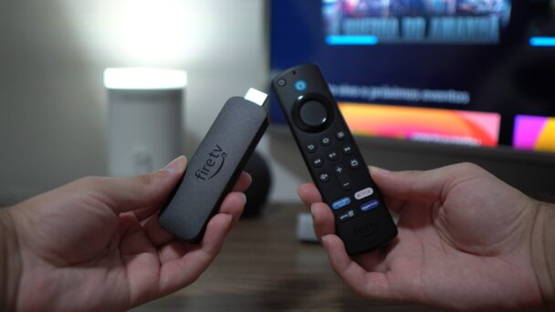 Two hands are holding a Fire TV Stick and its accompanying remote control against a blurred background that includes a television screen. The Fire TV Stick is in the left hand, and the remote control is in the right hand. - Cuponsy