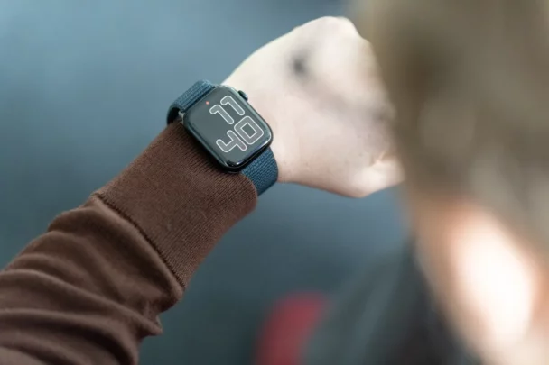 A person wearing a dark-colored smartwatch with a woven strap on their left wrist. The watch face displays the time as 11:40. The person is dressed in a long-sleeve brown shirt. The background is blurred, focusing on the watch and wrist. - Cuponsy