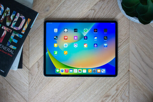 A tablet with a colorful home screen featuring various app icons on a wooden surface. Nearby is a book titled "MONDI LATE" with a colorful cover and part of a green succulent in a pot. - Cuponsy