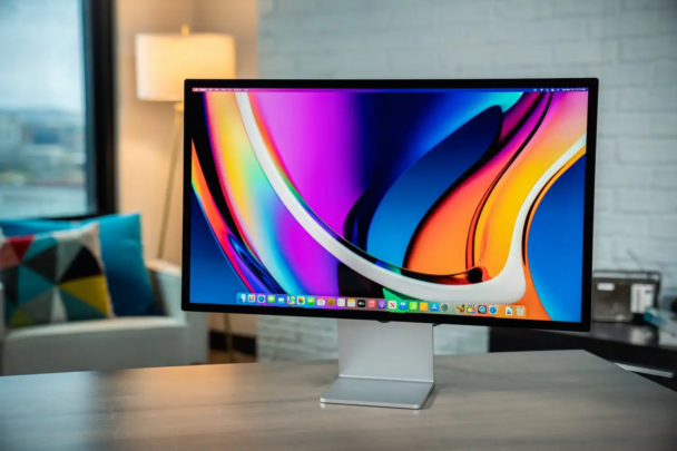 A sleek computer monitor displays a vibrant, abstract, colorful wallpaper. The monitor is on a stylish, minimalist desk in a modern office space with a geometric pillow on a white chair, a lit lamp, and a large window in the background. - Cuponsy