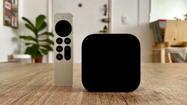 A sleek streaming device and its remote control sit on a wooden table in a modern home interior. The remote is white with a black circular control pad and buttons, while the device is a small, black, square-shaped box. A plant and bookshelves are visible in the background. - Cuponsy