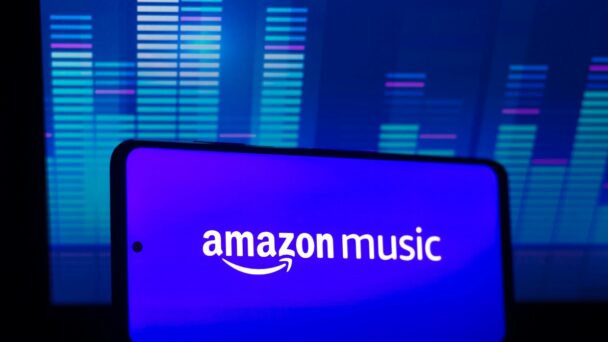 A smartphone is shown with the Amazon Music app open, displaying its logo prominently on the screen. In the background, there is a monitor with colorful sound wave patterns, suggesting audio activity or music visualization. The overall color scheme is blue and vibrant. - Cuponsy