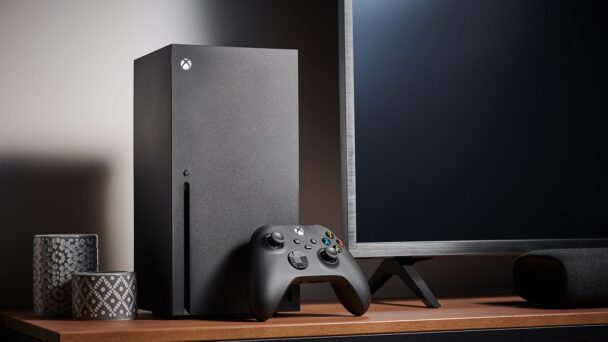 A sleek black gaming console and controller sit on a wooden desk beside a large monitor and decorative items. The setup is minimalistic, with the gaming console standing vertically and the controller placed in front of it. - Cuponsy