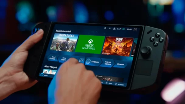 Close-up of a person holding a handheld gaming device displaying a screen with gaming options. The screen shows a list of recommended games, game settings, and an Xbox Game Pass icon. The person's finger is pointed at the screen, ready to make a selection. - Cuponsy