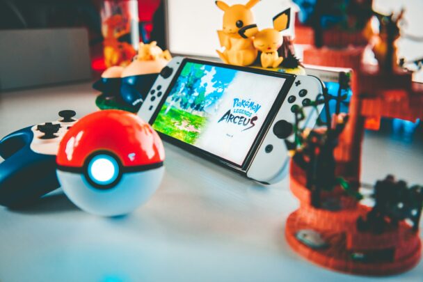 A Nintendo Switch with the game Pokémon Legends: Arceus on the screen sits on a white surface. Surrounding it are Pokémon-themed decorations, including a Poké Ball-shaped controller and Pikachu figures. The background shows more Pokémon figurines and displays. - Cuponsy