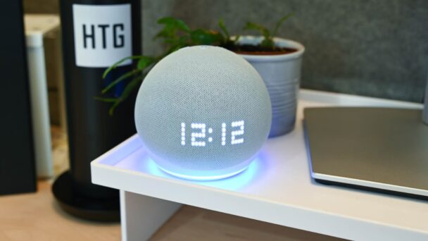 A smart speaker with a gray fabric exterior and digital clock display showing "13:12" sits on a white shelf. The base of the speaker emits a blue light. Next to it is a potted plant, and in the background, there is a black cylindrical item labeled "HTG. - Cuponsy