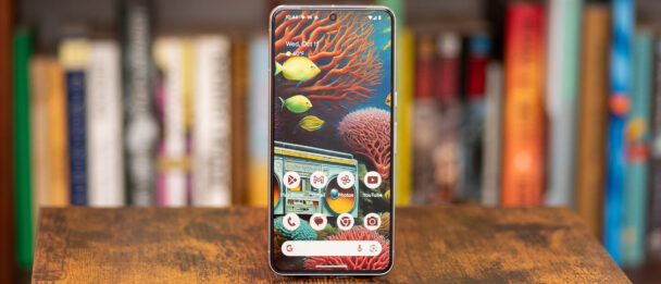 A smartphone with a colorful underwater-themed wallpaper is displayed on a wooden surface. The home screen shows various app icons including Gmail, Photos, YouTube, and Google. A bookshelf with assorted books is blurred out in the background. - Cuponsy