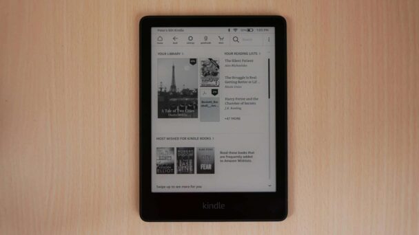 A black Kindle e-reader is placed on a light wooden surface, displaying the "Your Library," "Your Reading Lists," and "Most Wished For Kindle Books" sections on its screen. The title, "Here's my Kindle," is shown at the top of the device. - Cuponsy