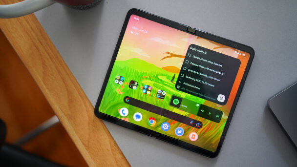 A foldable smartphone is partially unfolded on a gray desk. The screen displays a colorful landscape wallpaper and various app icons, with a to-do list widget and a music player widget featuring the Spotify app. A mug and a laptop are partly visible in the background. - Cuponsy