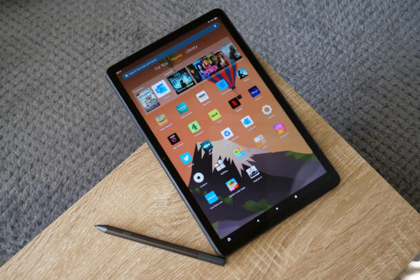 A black tablet with various app icons displayed on its screen rests on a wooden surface. A stylus lies diagonally in front of the tablet. The background features a carpeted floor. - Cuponsy