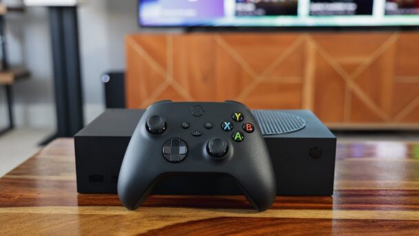 A black gaming console and controller rest on a wooden table. The console has a modern design with a vented circular section on top. The controller features the standard buttons: A, B, X, Y, a directional pad, and analog sticks. A cabinet is in the background. - Cuponsy