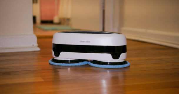 A Samsung robot mop cleaner is operating on a wooden floor. The device is primarily white with black accents and has two rotating blue cleaning pads underneath. The background shows a hallway with blurred details.