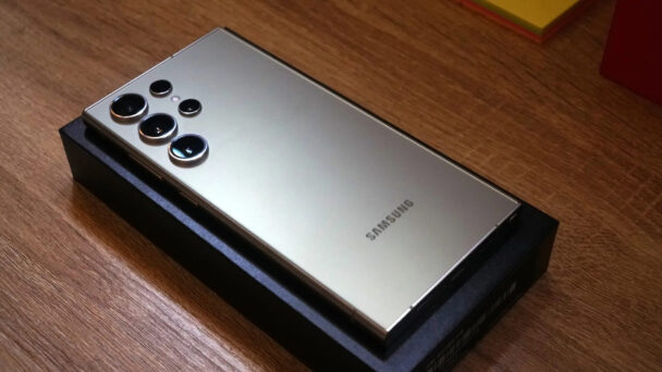 A gold Samsung smartphone with multiple camera lenses on the back rests on a black box, placed on a wooden surface. The phone has a sleek design, and the brand name "Samsung" is visible on the lower part of the back. - Cuponsy