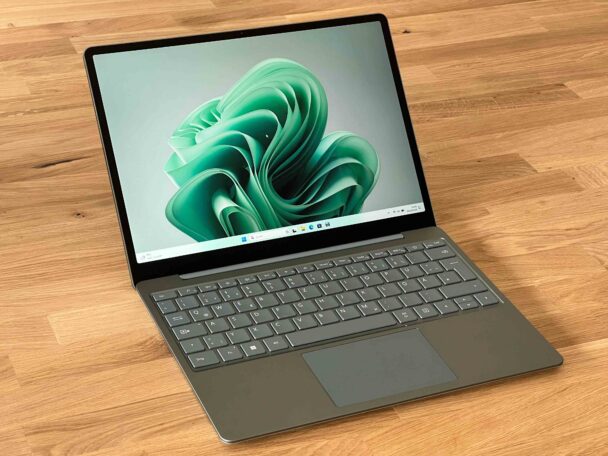 A laptop with a sleek gray design is placed on a wooden surface. The screen displays a green abstract wallpaper, and the taskbar at the bottom shows various icons and a search bar, suggesting it is running the Windows operating system.